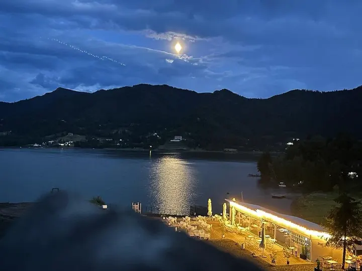 Fotografie de seară cu luna deasupra lacului și terasa luminată pe mal